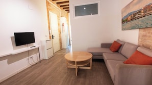Uma sala de estar moderna com televisão de tela plana, mesa de centro redonda de madeira, sofá cinza com almofadas laranjas e uma pintura de paisagem urbana na parede.