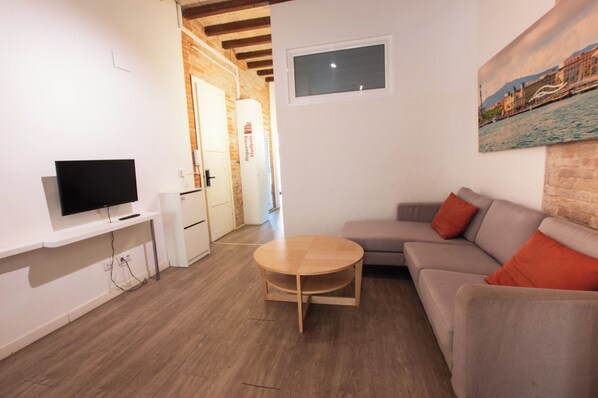 A modern living room with a flat-screen TV, a round wooden coffee table, a grey sofa with orange cushions, and a painting of a cityscape on the wall. - Classic Gracia Apartments (Barcelona)