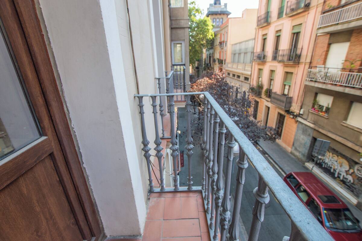 Ein schmaler Straßenblick von einem Balkon mit Holztür, Metallgeländer und einem roten Auto auf der Straße.