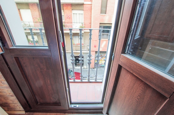 A view from a balcony with a wooden door and window, overlooking a street with a red car and a building with a balcony.