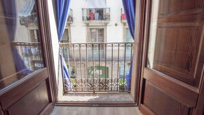 A view through a window with blue curtains, revealing a balcony with a metal railing and a building with balconies in the background. - Classic Catedral Apartments  (Barcelona)