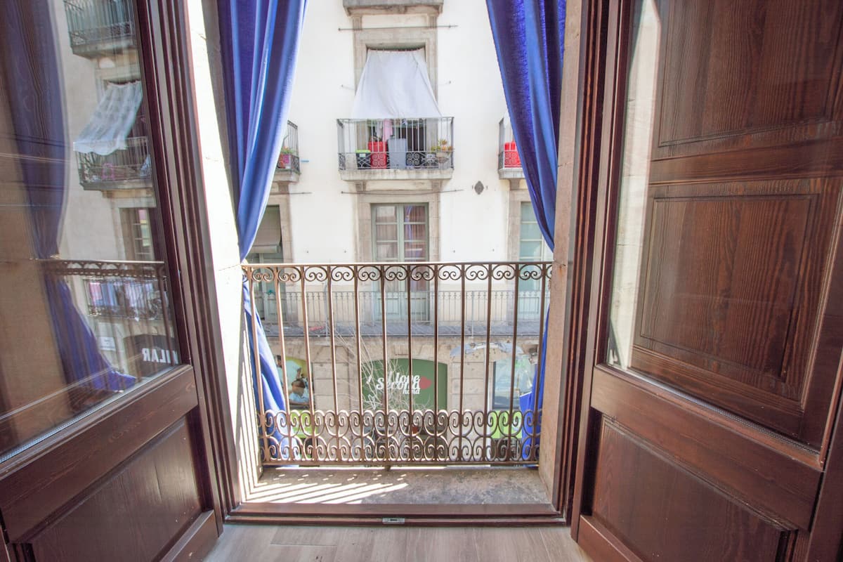 Blick durch ein Fenster mit blauen Vorhängen, der einen Balkon mit Metallgeländer und ein Gebäude mit Balkonen im Hintergrund zeigt.
