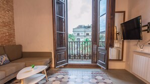 A living room with a sofa, a coffee table, a TV mounted on the wall, and a balcony with a view of buildings. - Barcelona Classic Ramblas - Plaza Real (Barcelona)