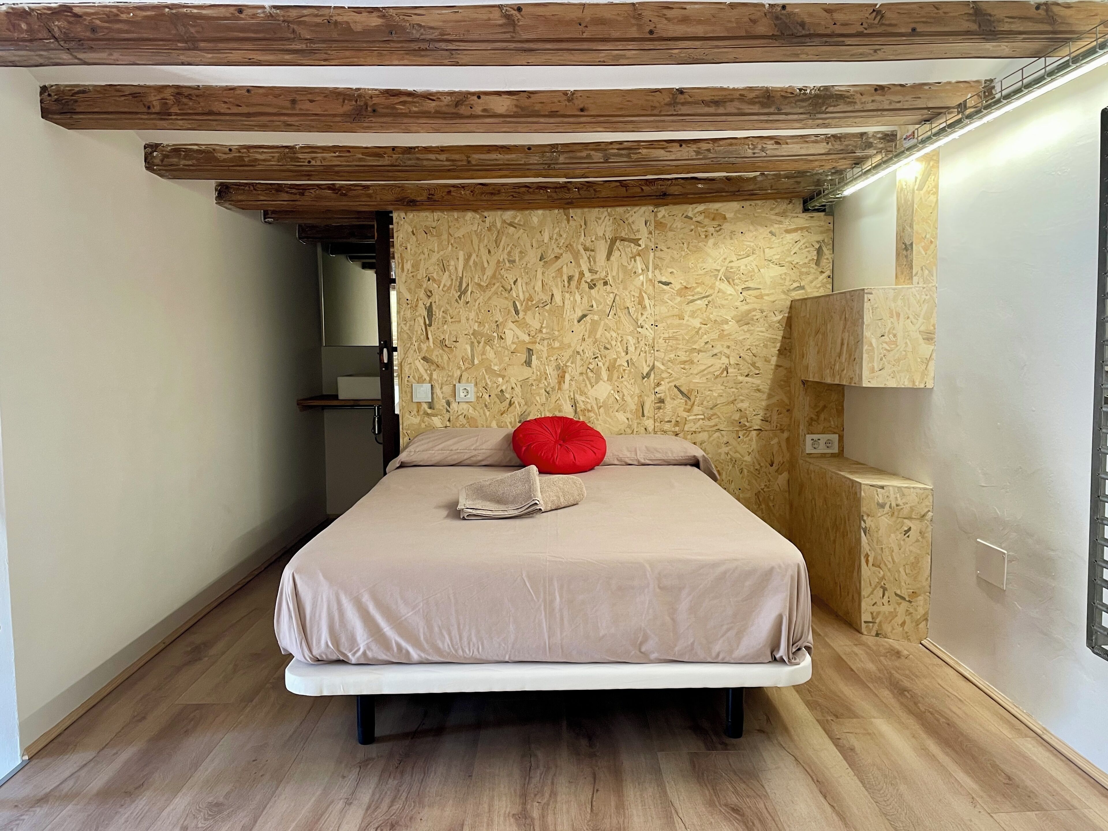 Ein Schlafzimmer mit Holzboden, einem Bett mit beiger Bettdecke und rotem Kopfkissen, einer Holzwand und einer kleinen Regaleinheit.