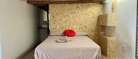 Ein Schlafzimmer mit Holzboden, einem Bett mit beiger Bettdecke und rotem Kopfkissen, einer Holzwand und einer kleinen Regaleinheit.