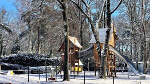 Un patio de juegos cubierto de nieve con un tobogán y estructuras para escalar, rodeado de árboles.
