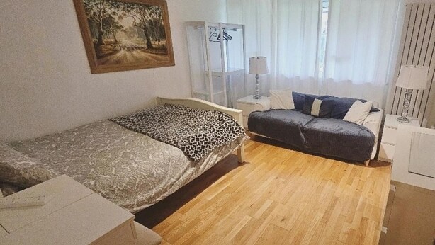 Una stanza piccola e funzionale con un letto, una scrivania e una sedia. Si vede una finestra con tende, una lampada e un quadro appeso al muro.