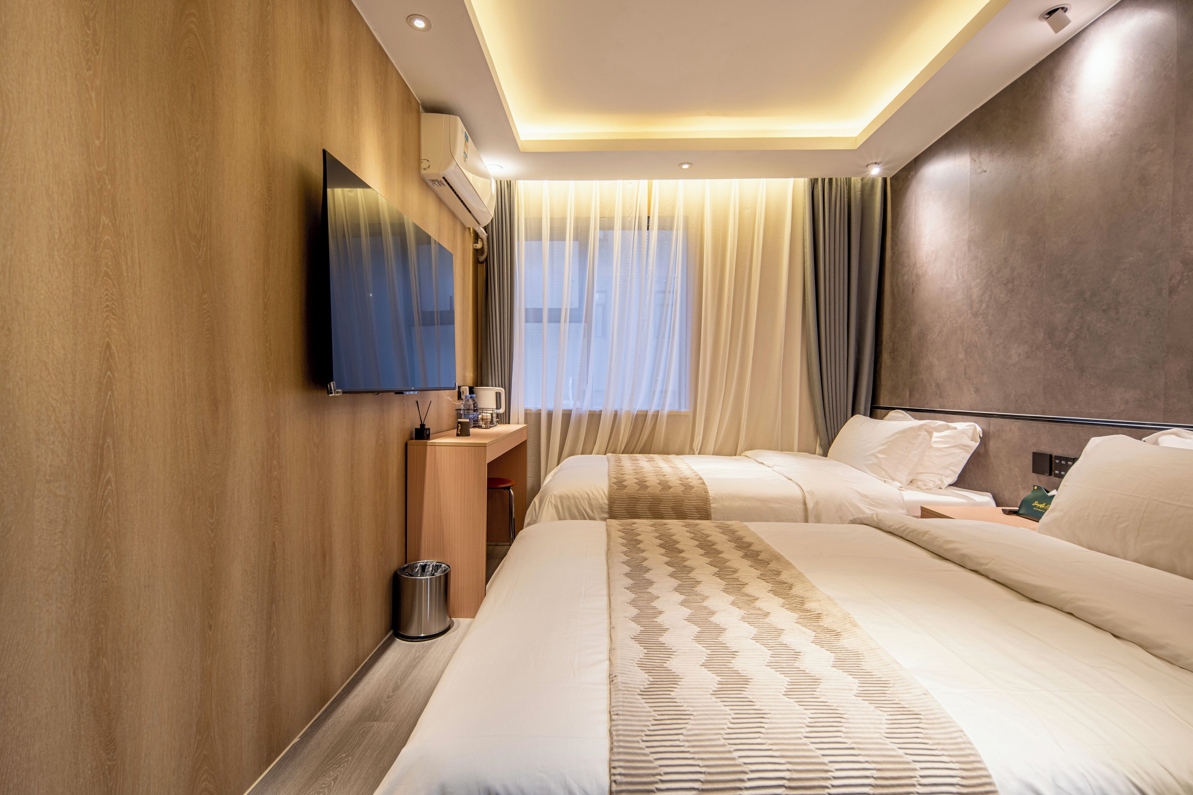 Habitación de hotel con dos camas, un televisor montado en la pared, un escritorio con una silla y una ventana con cortinas.