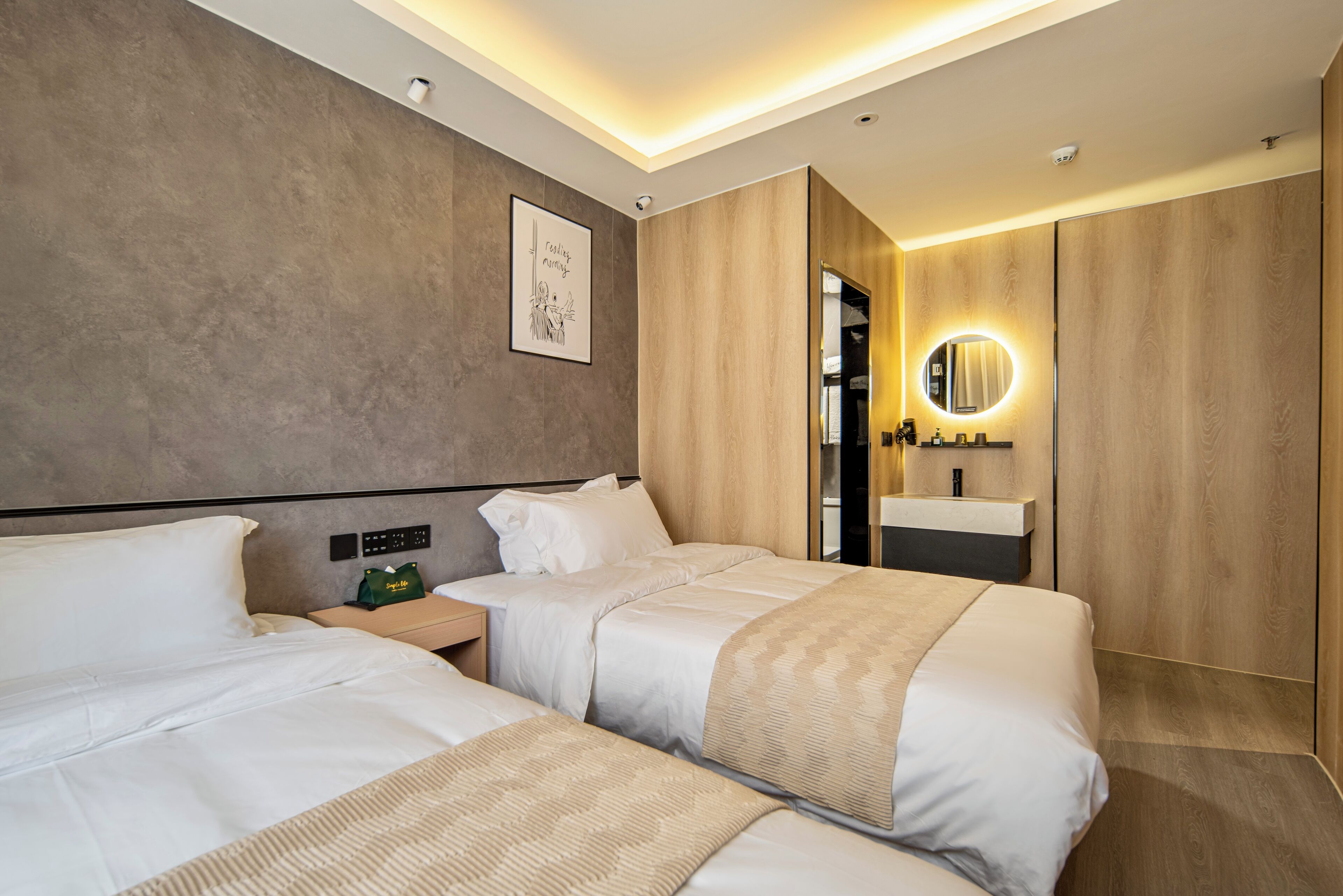 A modern hotel room with two beds, a bedside table with a lamp, a wall-mounted mirror, and a framed picture.