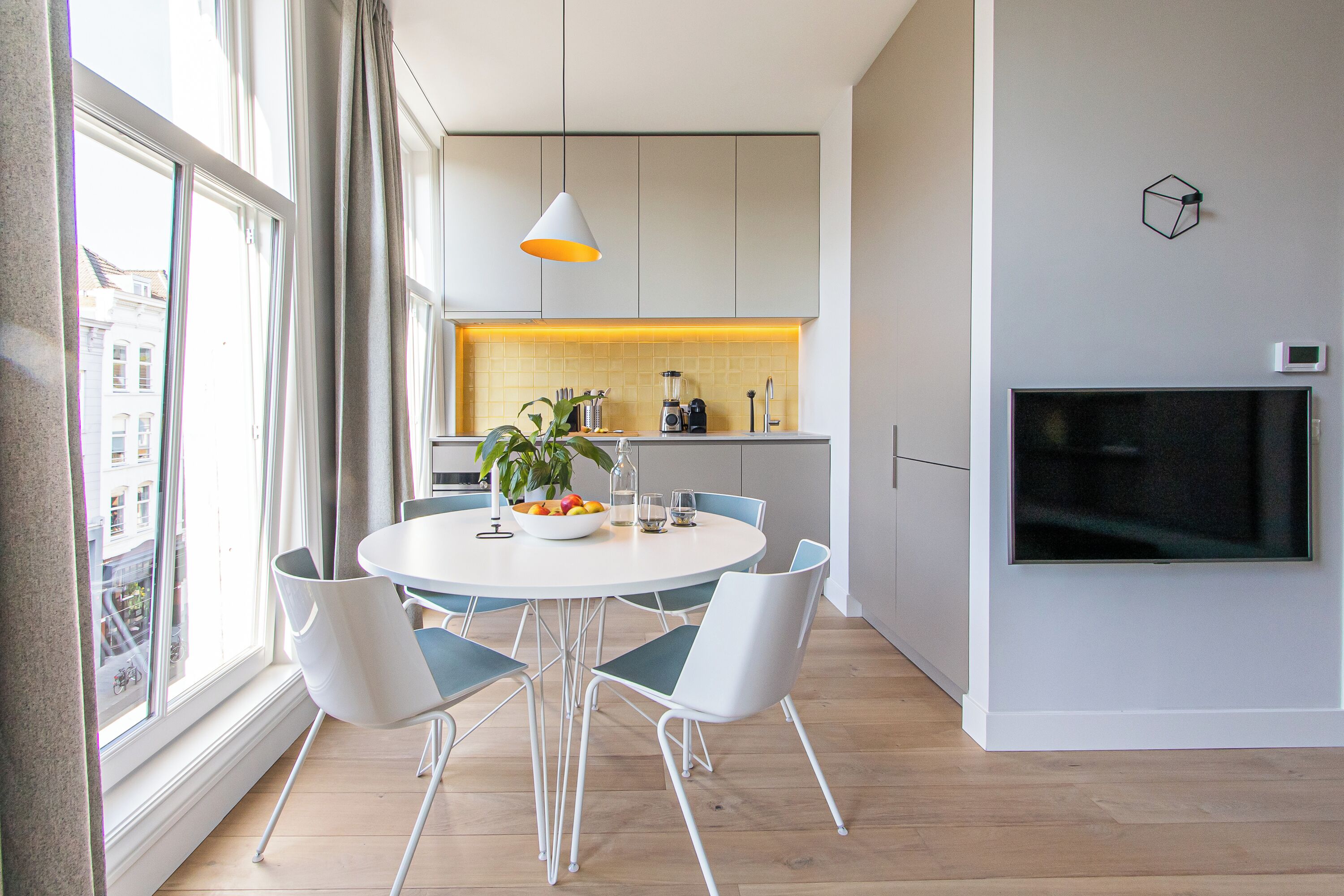 Una cocina moderna con armarios blancos, una mesa de comedor con cuatro sillas, una planta y un televisor montado en la pared.