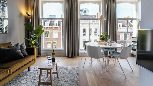 A modern living room with a sofa, dining table, chairs, and large windows with curtains. - City Apartment Darling (Rotterdam)