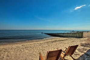 A beach with two wooden chairs facing the sea, a wooden pier extending into the water, and a clear blue sky.