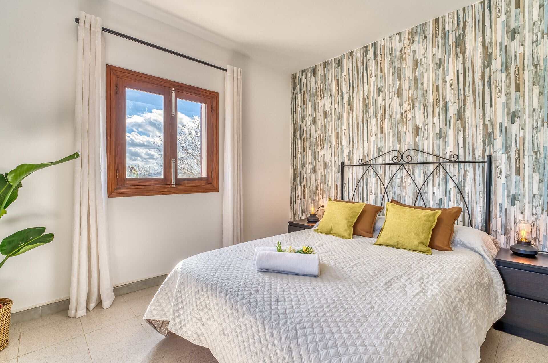 Ein Schlafzimmer mit einem großen Bett, einem Fenster mit Vorhängen, einer Wand mit vertikalen Holzlatten und einem Nachttisch mit einer Lampe.