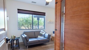 A room with a gray sofa, a small side table, a window with blinds, and a wooden door. - Cozy Dark Sky Retreat Near Sedona • Hot Tub, Sauna & Views (Cornville)