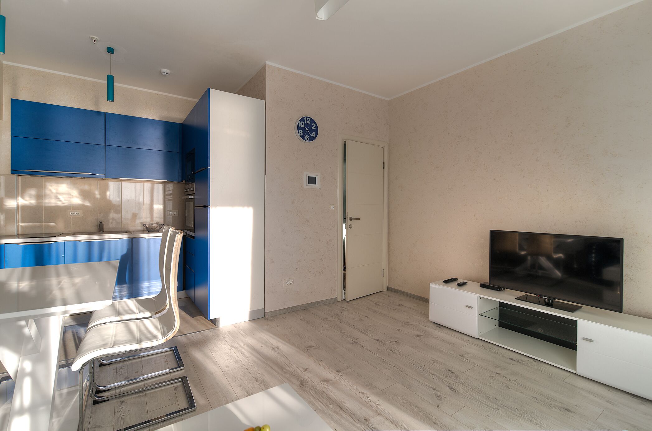 A modern living room with a flat-screen TV, a dining area with blue cabinets, and a kitchen with white cabinets.