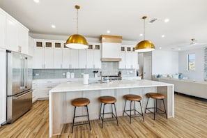 Una cucina moderna con mobili bianchi, un frigorifero in acciaio inox, una grande isola con sgabelli e lampade a sospensione dorate.
