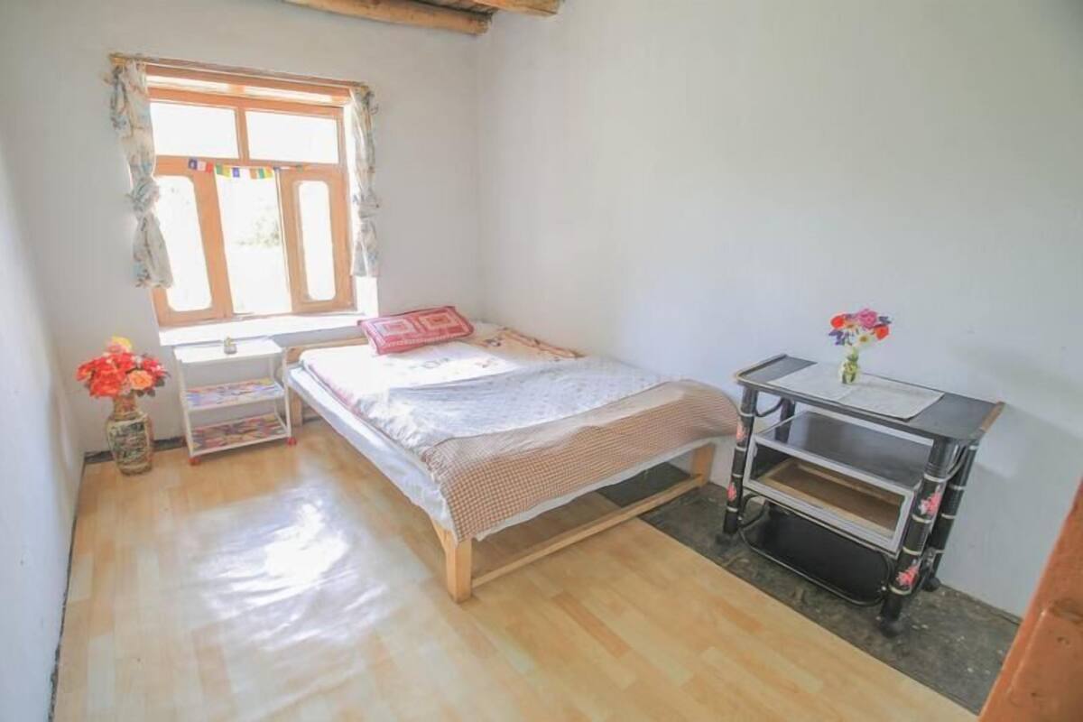 A small, single-bed room with a wooden floor, a window with curtains, a bedside table with a vase of flowers, and a desk with a chair.