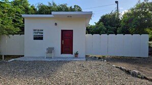 A small, single-story building with a red door, a white picket fence, and a gravel driveway. - Sunburst Studio (Caicos Islands)