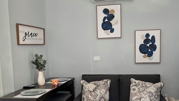 A modern living room with a dark grey sofa, two patterned throw pillows, a small side table with a vase and candle, and a framed inspirational quote on the wall. - Sunburst Studio (Caicos Islands)