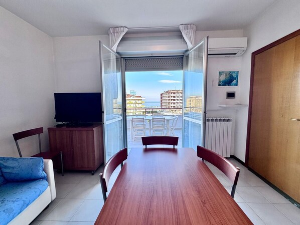 A room with a wooden dining table, a TV on a cabinet, a sofa, and a balcony with a view of buildings.