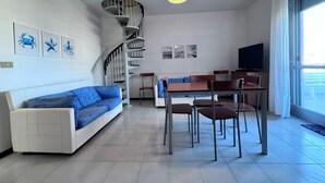 A modern living room with a white sofa, blue cushions, a wooden dining table with chairs, a TV, and a spiral staircase. - Apartment with all amenities 50 meters from the sea. Breathtaking view (Lido Riccio)