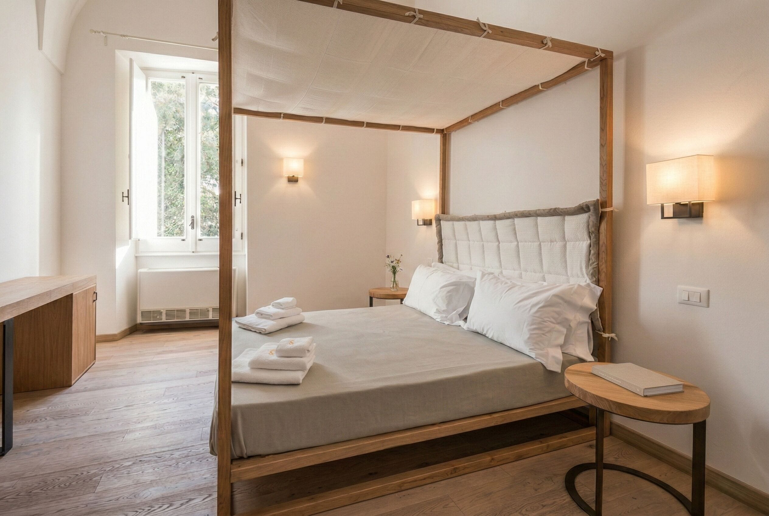 A four-poster bed with white linens, a bedside table with a book, and two wall-mounted lamps.