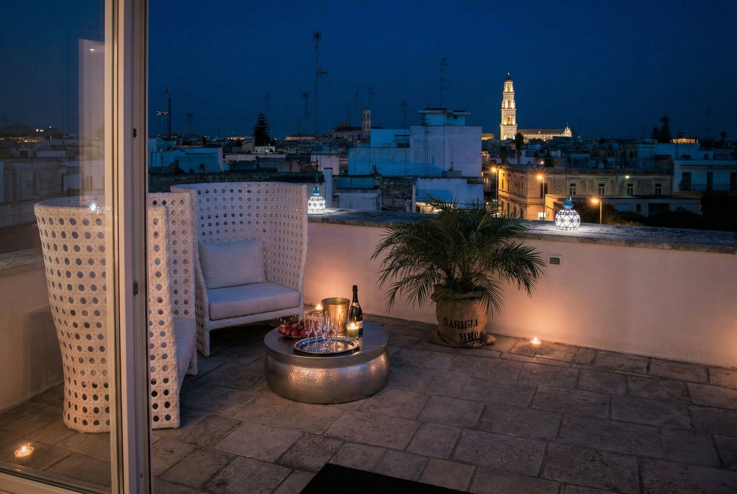 A rooftop terrace with a seating area, a potted plant, and a table with a bottle and glasses.
