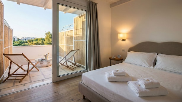 Een hotelkamer met een bed, nachtkastje, lamp, uitzicht door een schuifdeur, en een balkon met stoel en tafel.