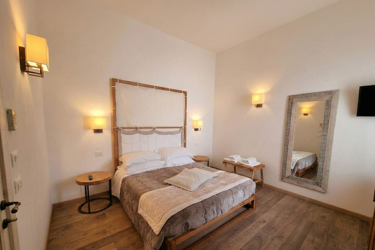 Un dormitorio con cama de madera, mesitas de noche, un espejo y luces fijadas en la pared.