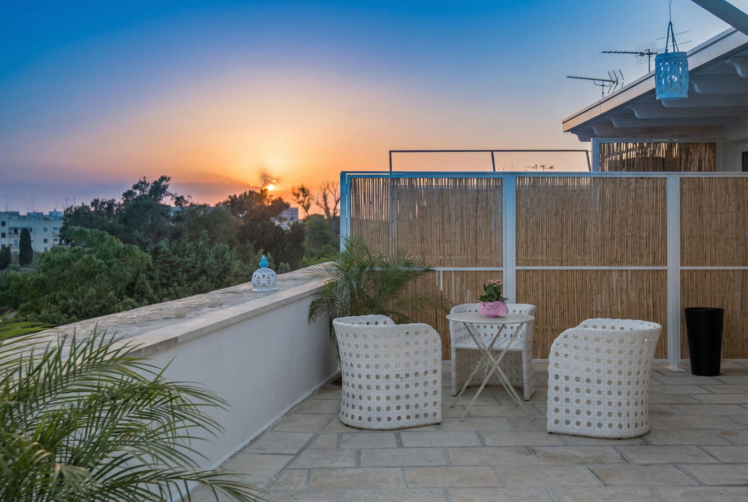 A rooftop terrace with a view of the sunset, a bamboo fence, and outdoor furniture.