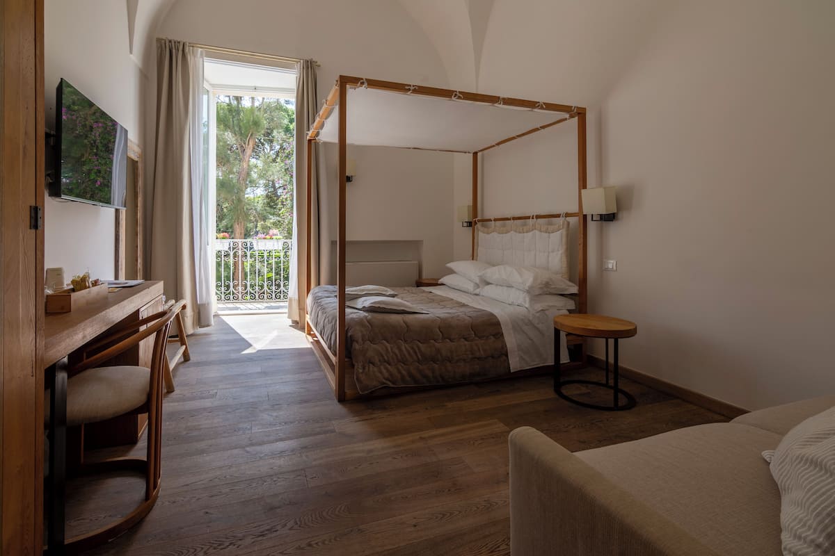 Un dormitorio con cama con dosel, muebles de madera, un escritorio con silla y un balcón con vistas a árboles.
