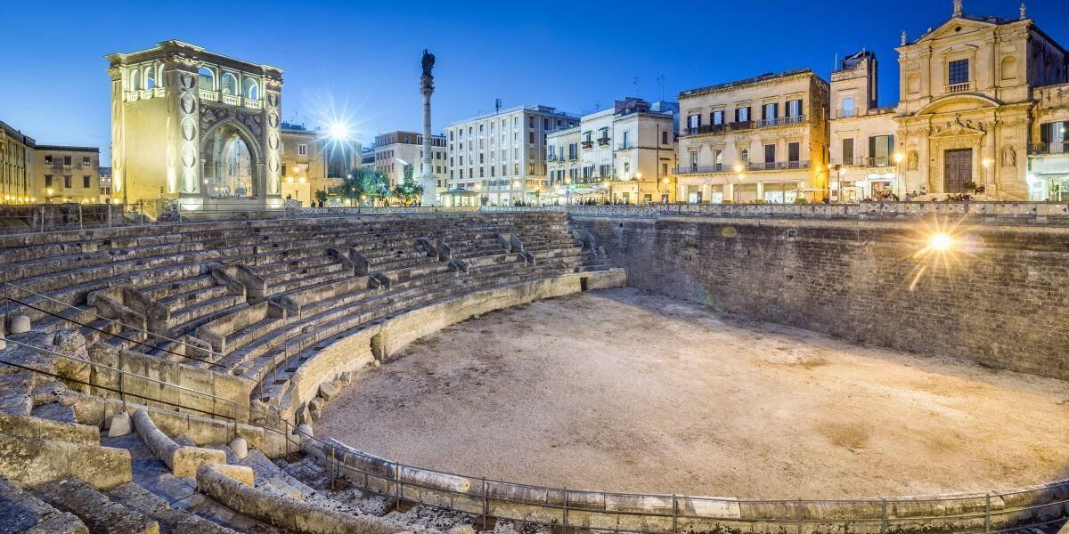 An ancient amphitheater with stone steps and a central stage, surrounded by historic buildings.