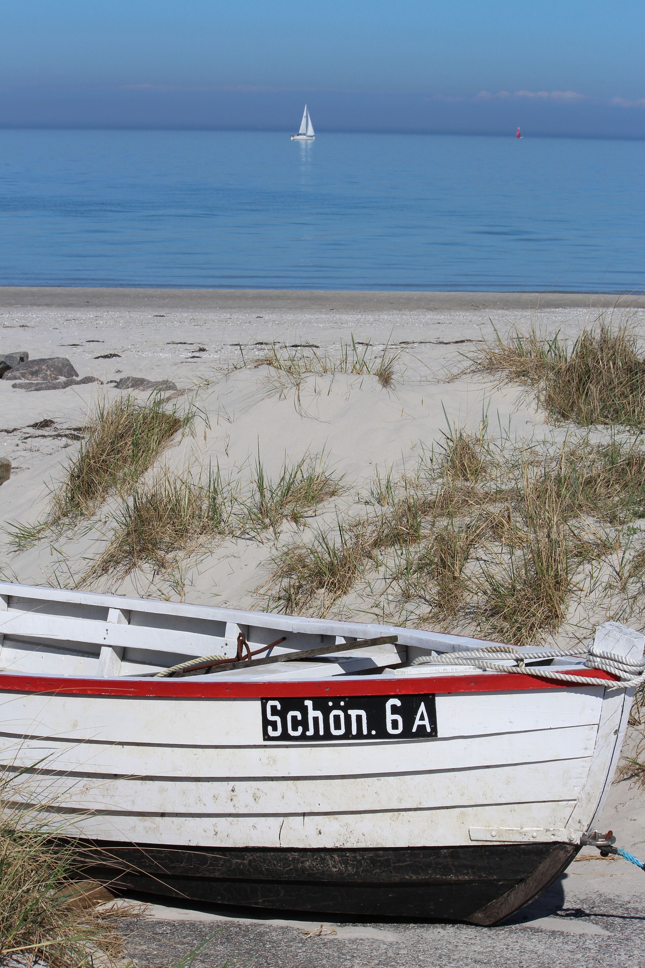 Ein Boot namens "Schön. 6A" am sandigen Strand, mit Gras und ruhiger See im Hintergrund.