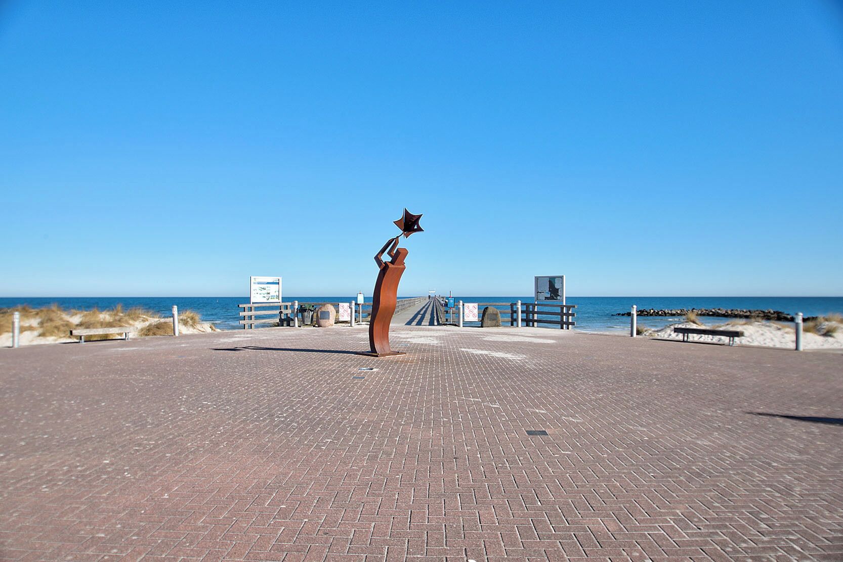 Ein gepflasterter Weg führt zu einem Strand mit einer Skulptur, Bänken und Informationstafeln.
