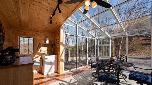 A wooden cabin with a dining area featuring a glass enclosure, a ceiling fan, and a kitchen with a stove and sink.