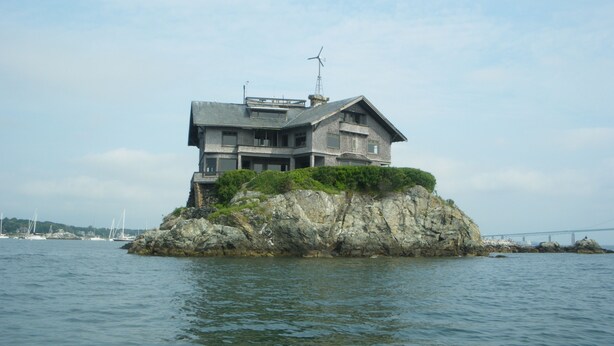 Ein modernes Haus auf einer felsigen Insel mit Blick auf das Meer und einer Brücke im Hintergrund.