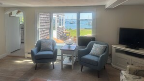 Ein Wohnzimmer mit zwei Sesseln, einem kleinen Tisch und einem Fernseher auf einem weißen Schrank. Große Fenster bieten Blick auf einen Steg und Boote.