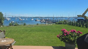 Eine Terrasse mit Tisch und Stühlen, ein Blumentopf mit rosa Blüten sowie der Blick auf eine Marina mit Booten und einer Brücke im Hintergrund.