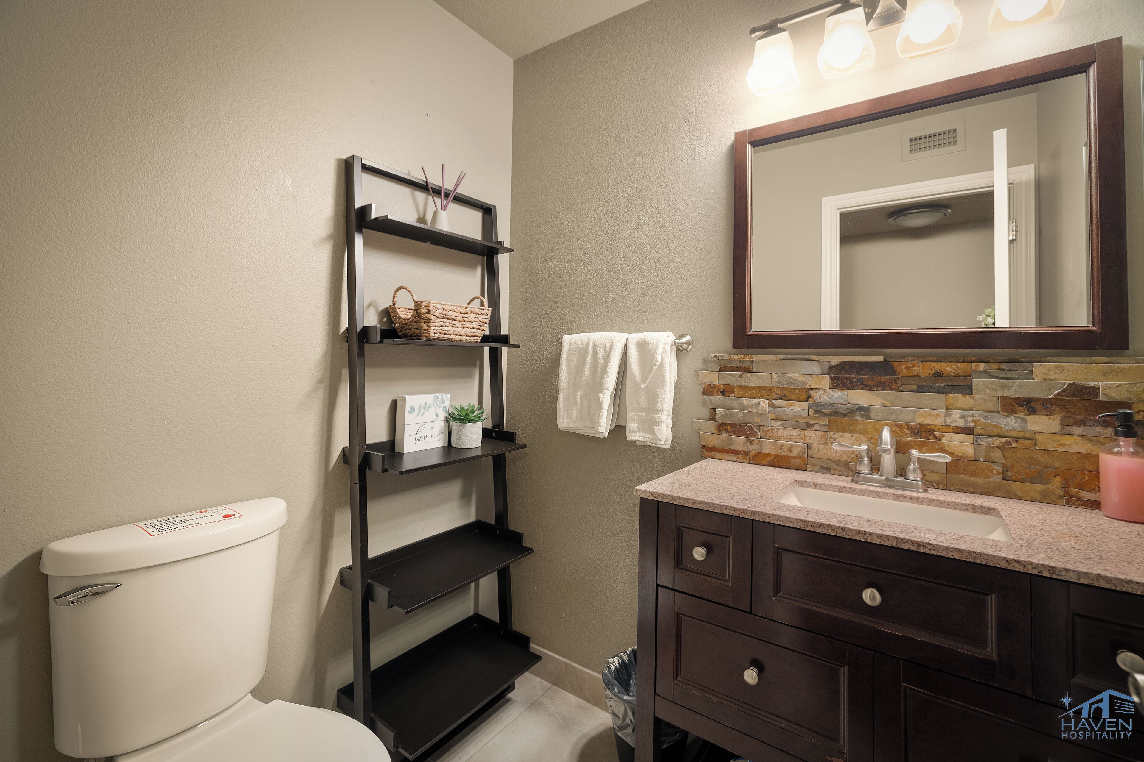 A bathroom with a toilet, a vanity with a sink, a mirror, and a shelf with toiletries.