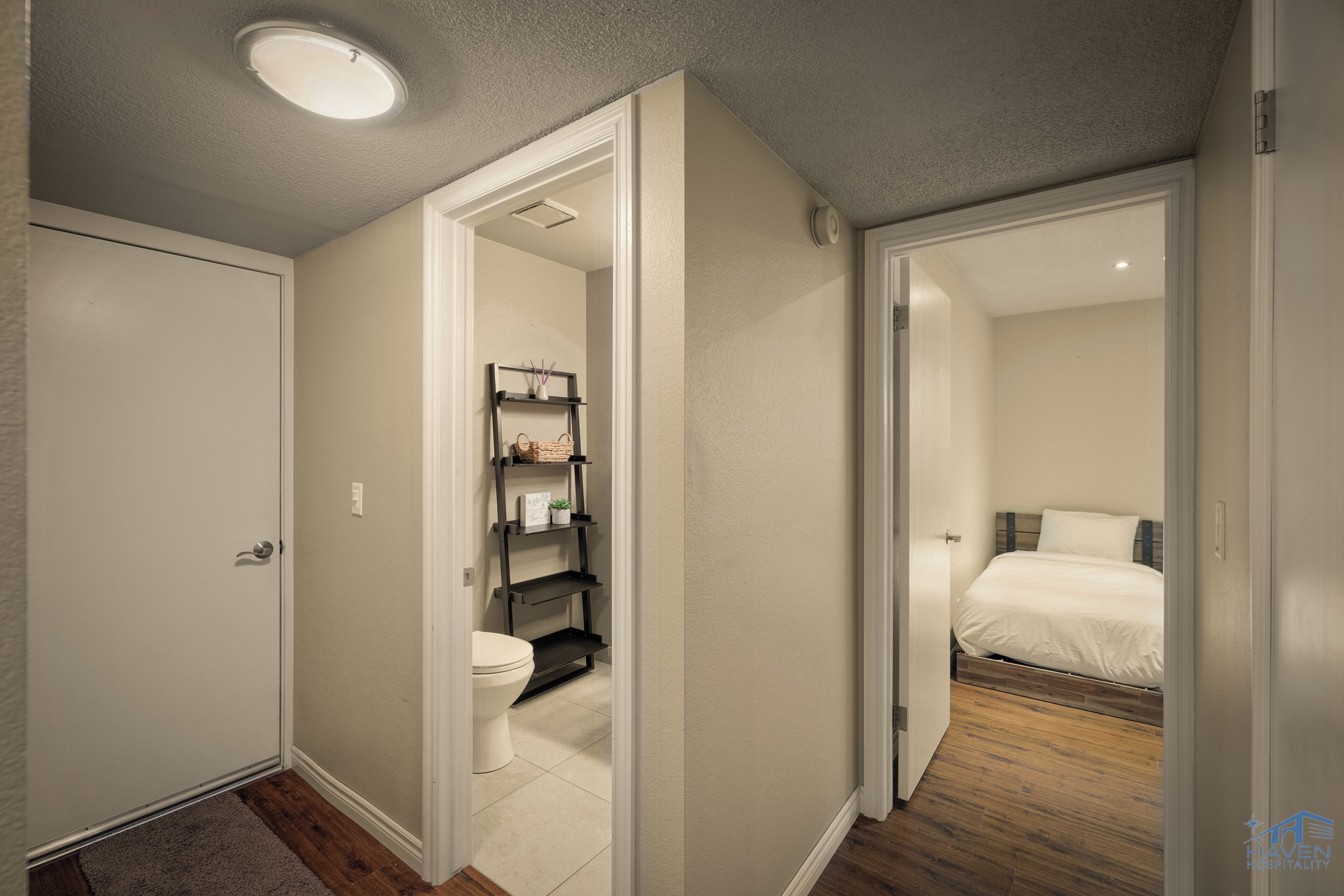 A narrow hallway leading to a bathroom with a toilet and shelves, and a bedroom with a bed and wooden flooring.