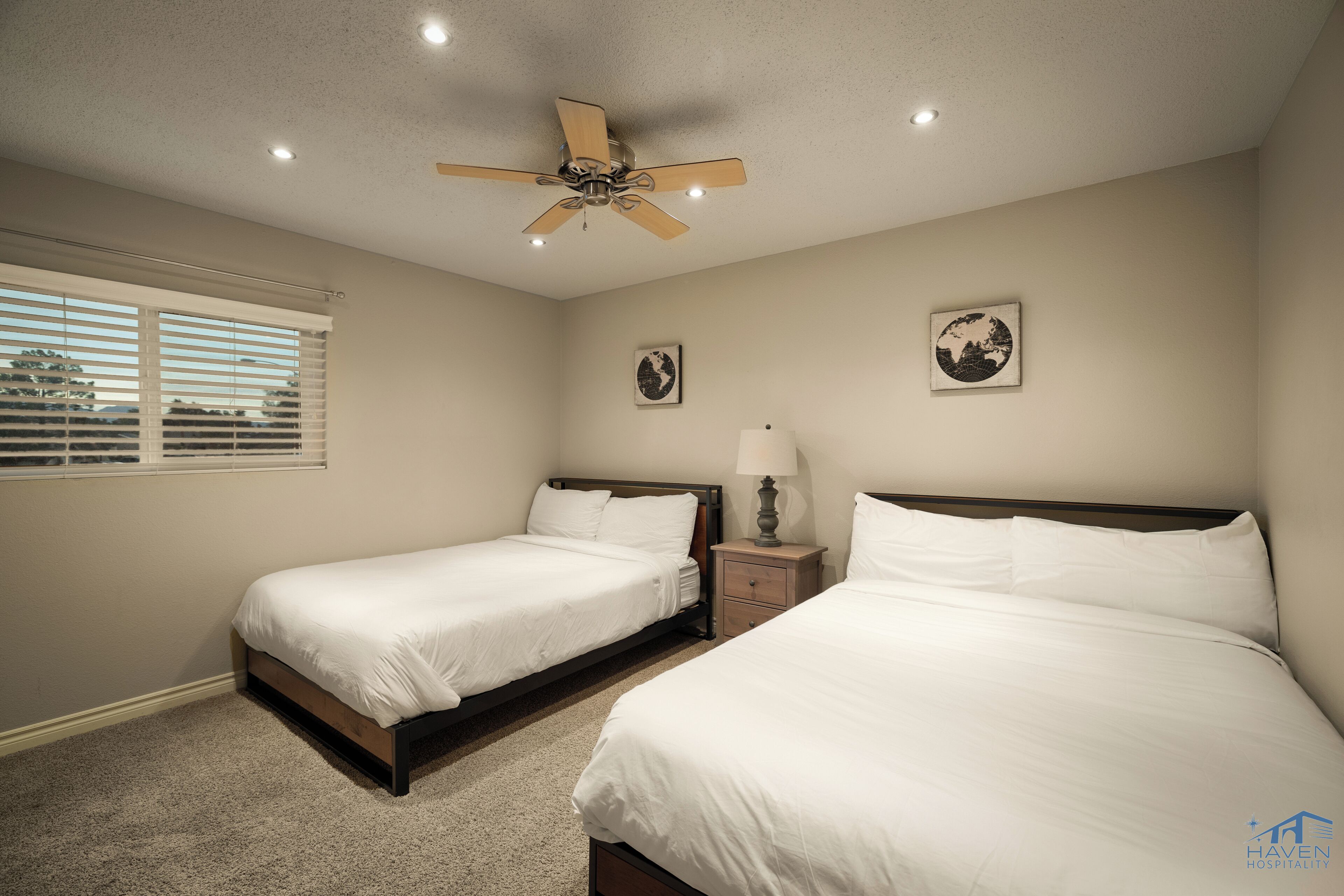A hotel room with two beds, a ceiling fan, a window with blinds, and two wall-mounted pictures.