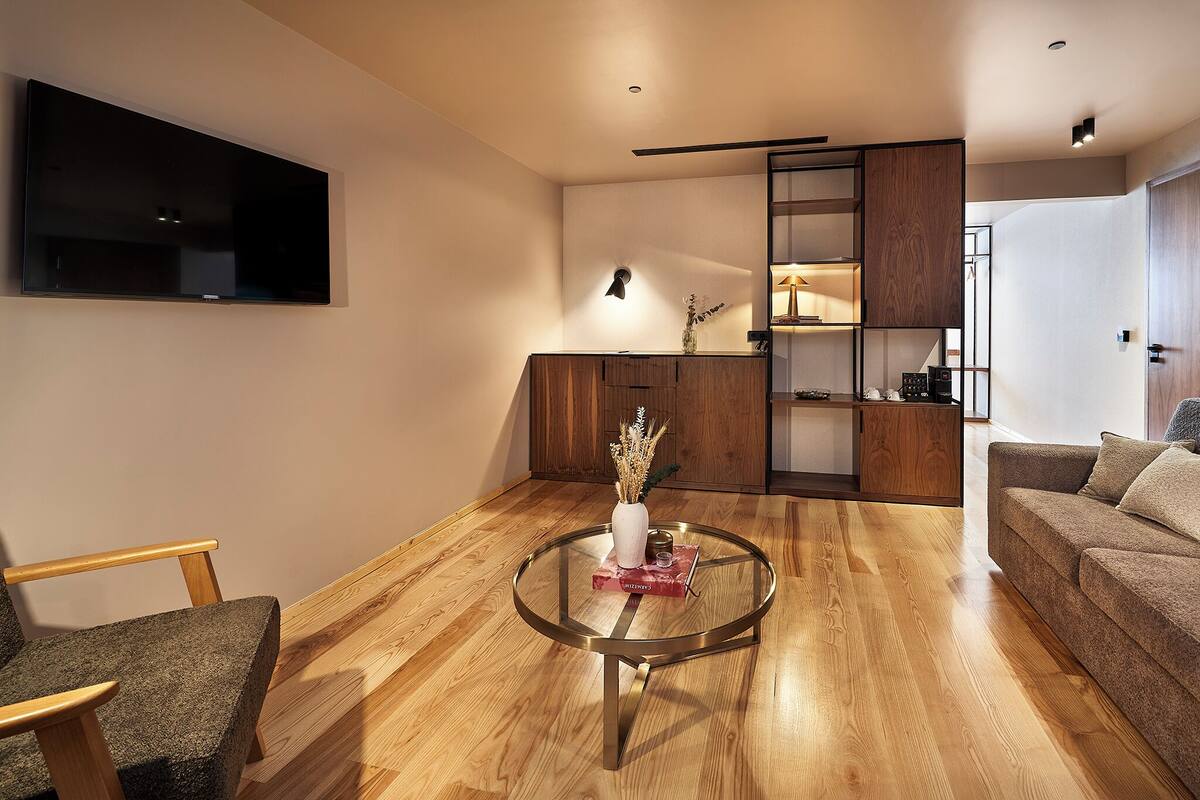 A modern living room with a round glass coffee table, wooden flooring, a flat-screen TV mounted on the wall, and a sofa.