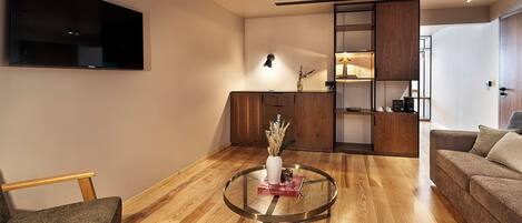 A modern living room with a round glass coffee table, wooden flooring, a flat-screen TV mounted on the wall, and a sofa.