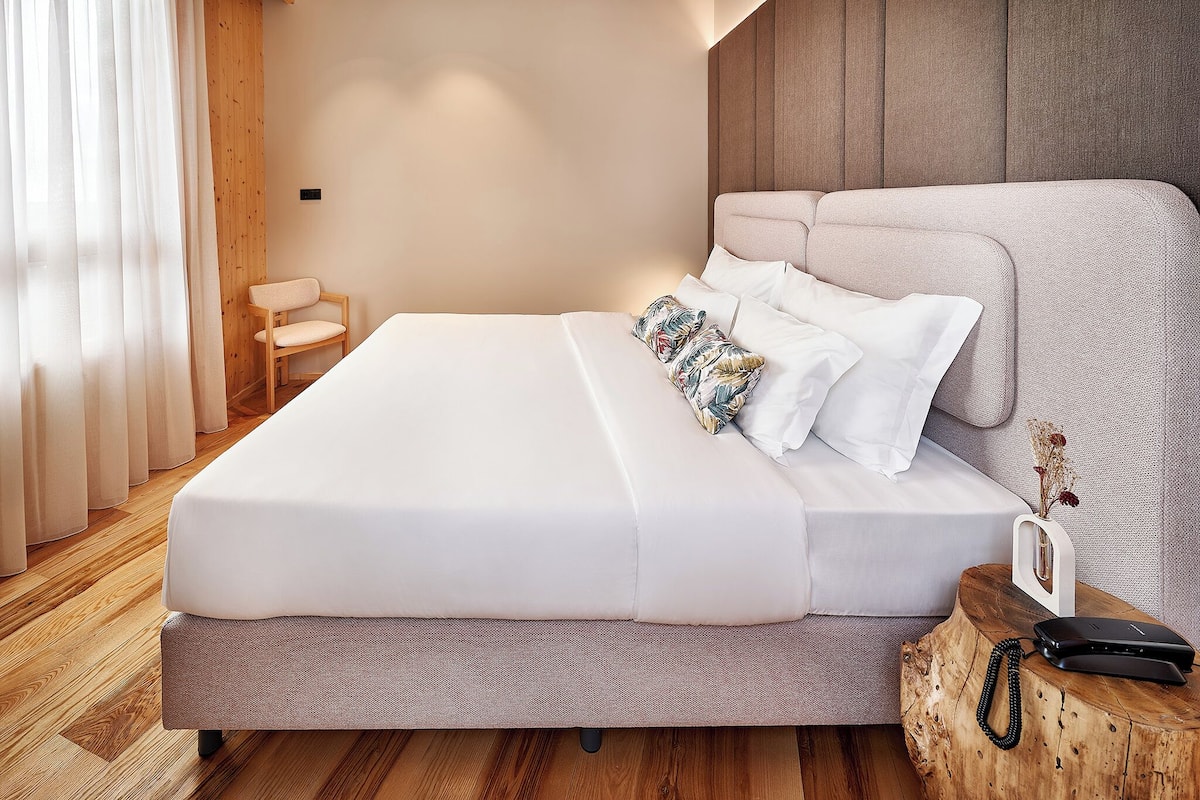 A neatly made bed with white linens, a wooden nightstand with a phone and a small vase, and a wooden chair in a modern bedroom with wooden flooring and a wooden headboard.
