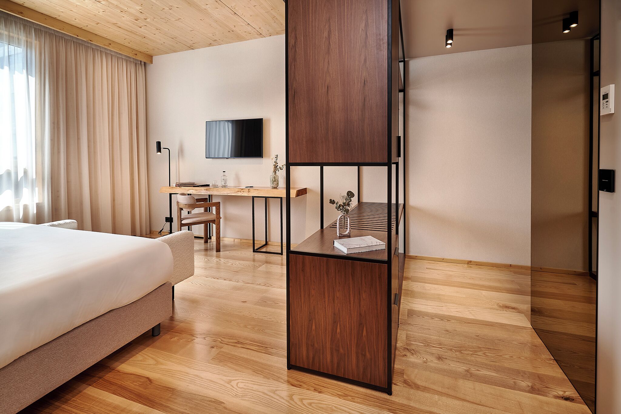 A modern hotel room with a large bed, wooden flooring, a desk with a chair, a television, and a decorative shelf.