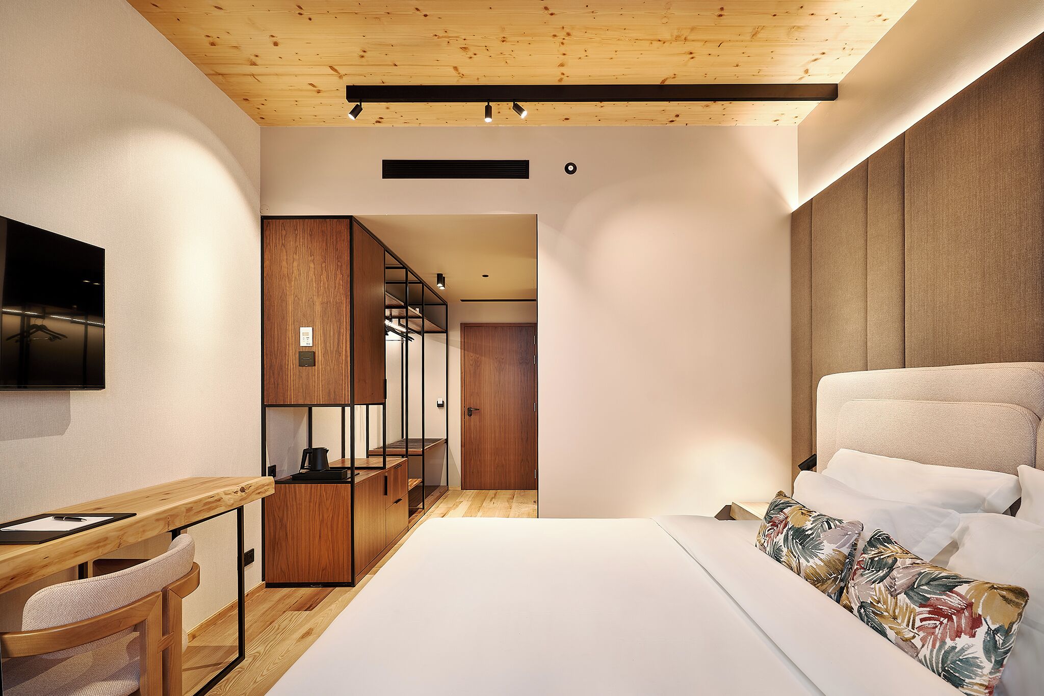 Un dormitorio moderno con techo de madera, cama con ropa de cama blanca, escritorio con silla y una zona de cocina con armarios de madera.