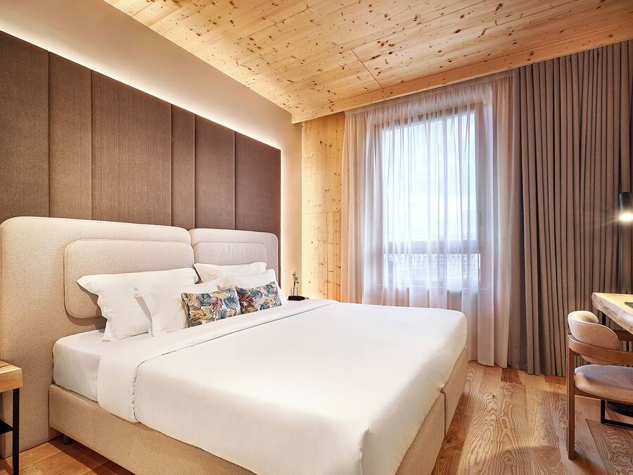 A modern hotel room with a large bed, a wooden ceiling, and a view of the outdoors through sheer curtains.