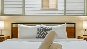 Un lit bien fait, avec du linge de lit blanc, un coussin à motifs et une serviette enroulée. Deux lampes de chevet sont posées sur des tables de nuit en bois.