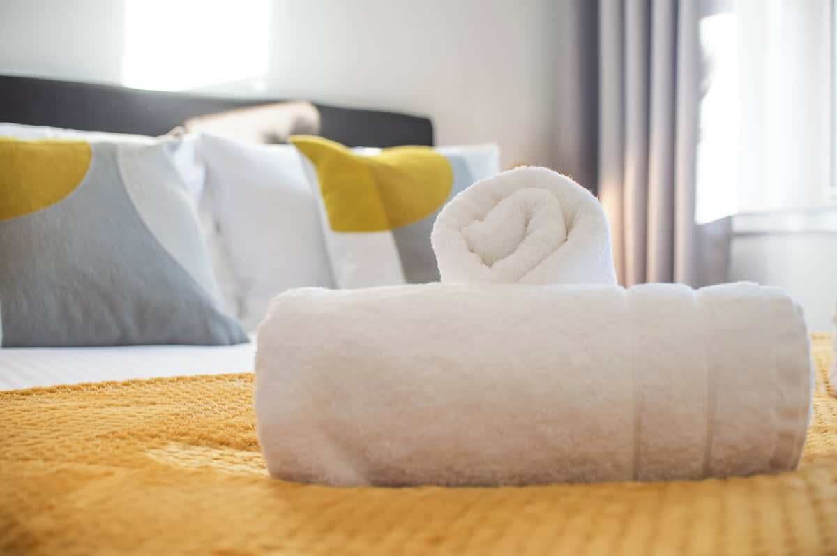 A neatly made white towel with a heart design on a bed with a yellow blanket, white pillows, and a grey pillow with a yellow accent.