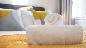 A neatly made white towel with a heart design on a bed with a yellow blanket, white pillows, and a grey pillow with a yellow accent.